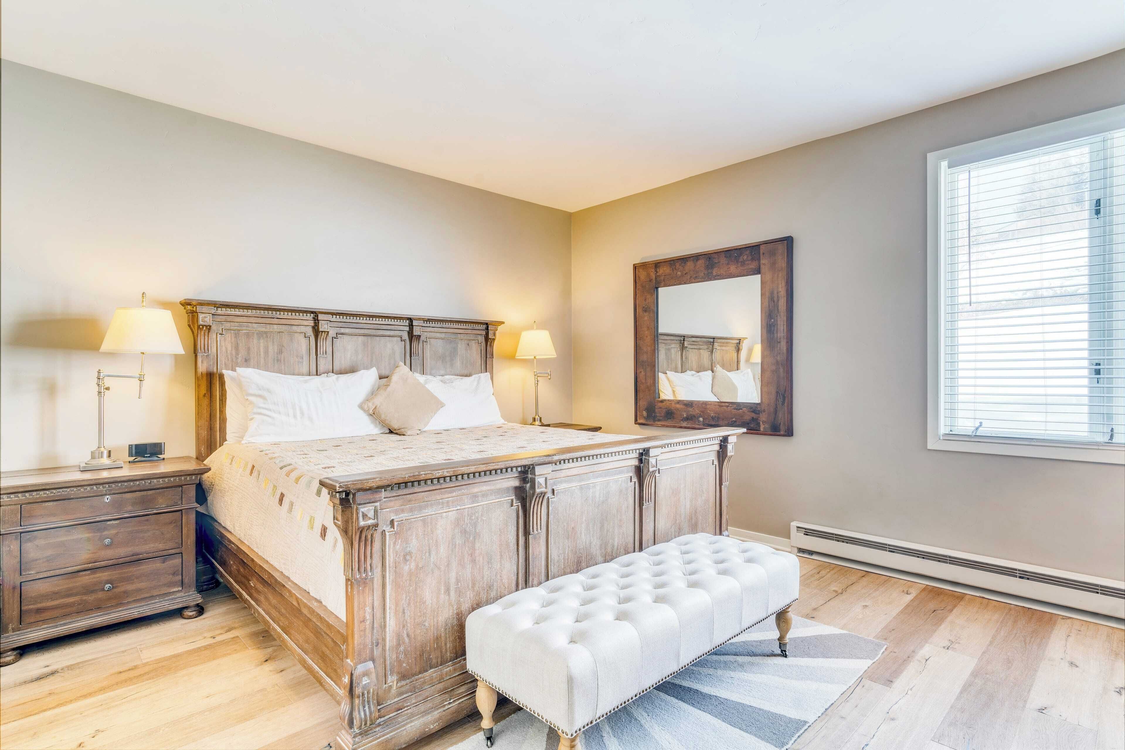 A bedroom with a wooden bed, bedside tables, a bench, a mirror, and a window.