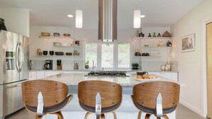 A modern kitchen with a central island, stainless steel appliances, wooden chairs, pendant lights, and a view of greenery outside. - Secluded + Modern Enclosed Patio, Forest Views Woodland Zen by Avantstay (Housatonic)