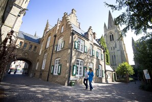 En historisk bygning med murstensfacade og grønne skodder, en brostensbelagt gade og en kirke i baggrunden.