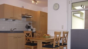 A compact kitchen with wooden cabinets, a built-in stove, and a dining table with four chairs. A clock is mounted on the wall, and a window with blinds is visible. - 1 in House Weser 1 Comfortable Vacation Home (Cuxhaven)