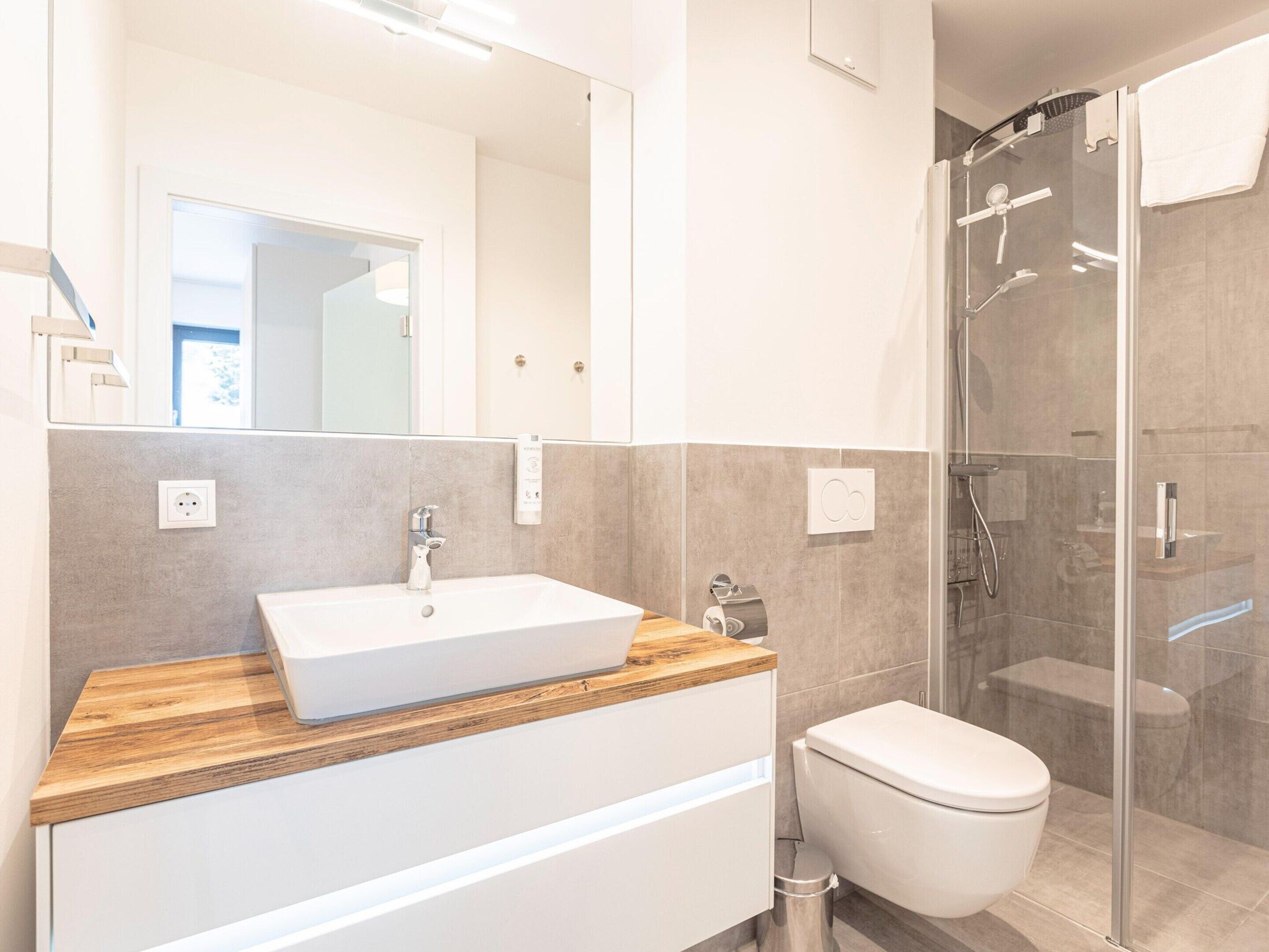 Une salle de bain moderne avec un meuble-lavabo en bois, un lavabo blanc, des toilettes et une douche séparée par une paroi en verre.