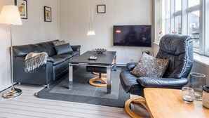 A modern living room with a leather sofa, a coffee table, a wooden chair, a TV, and a large window. - 4 Person Holiday Home in Blavand (Blavand)