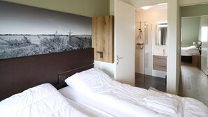 A hotel room with a bed, a wooden headboard, a large wall-mounted picture, a shower area, and a bathroom with a mirror.