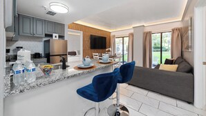 Une cuisine moderne dotée de plans de travail en granit, d’un four encastré et d’un téléviseur à écran plat. On y trouve également un coin repas avec un canapé gris et une table basse.