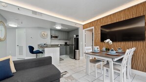 Une cuisine moderne avec des meubles gris, un four à micro-ondes et un réfrigérateur. On y trouve un coin repas avec une table et des chaises, et un canapé gris au premier plan.