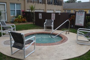 Ein kleiner Pool im Freien mit Ziegelkanten, umgeben von einer Betonplattform. Vier weiße Stühle mit Armlehnen und eine Metallleiter zum Pool sind vorhanden. Ein Schild weist darauf hin, dass es sich um Privatgrundstück handelt und Spa-Regeln gelten.