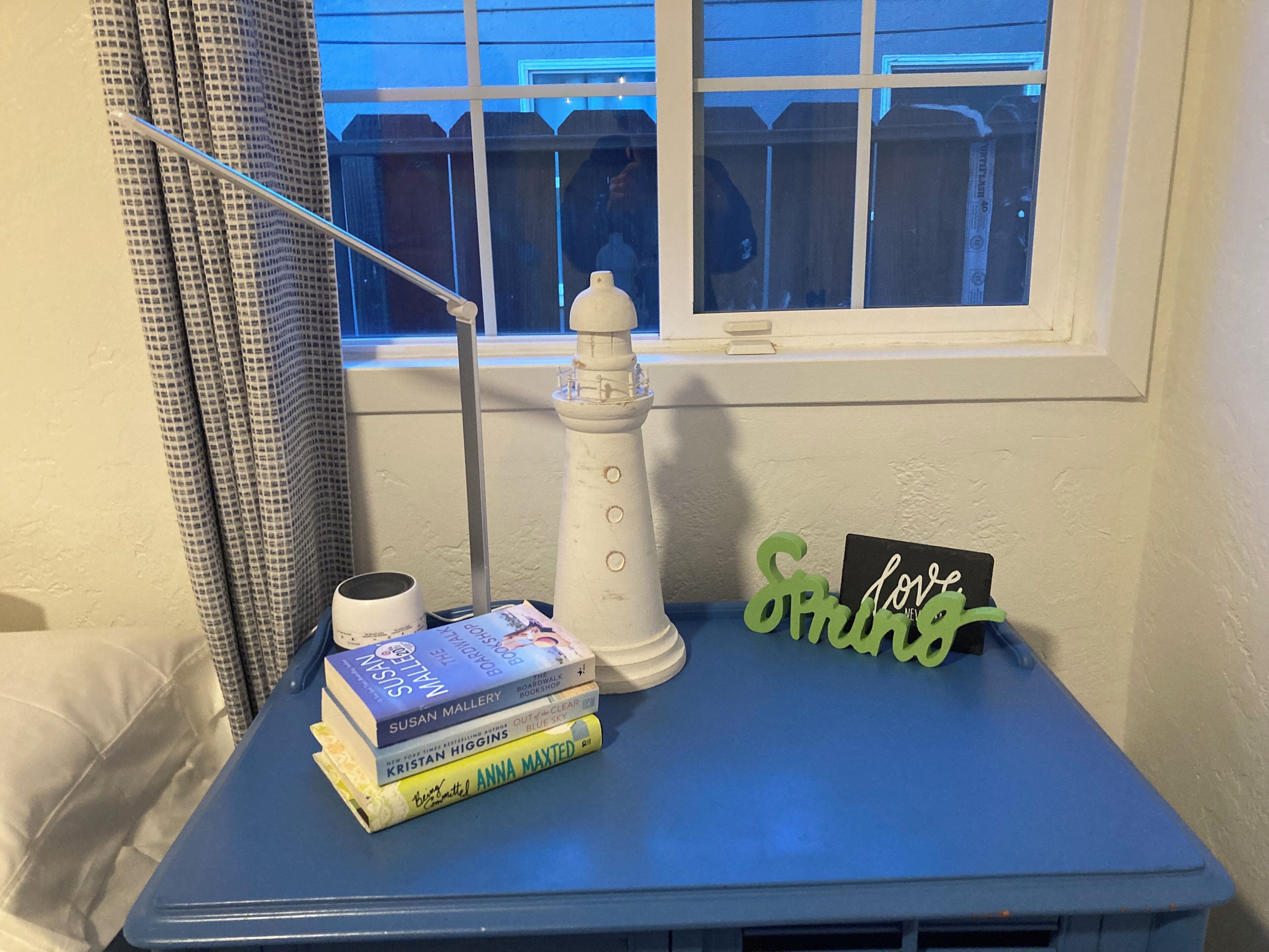 A small table with a stack of books, a white lighthouse model, a green sign, and a lamp.