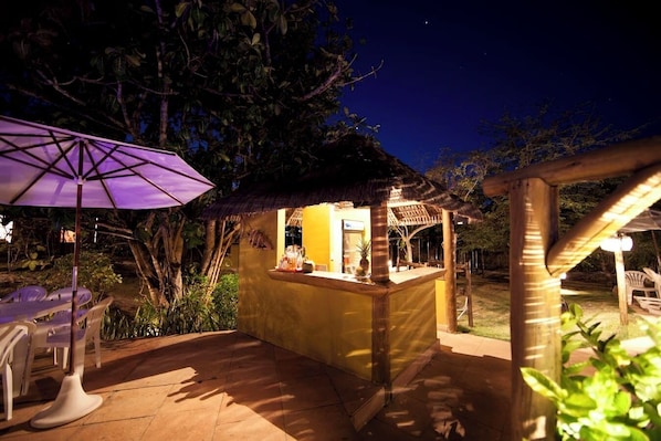 Un área de bar al aire libre con techo de paja, rodeada de follaje y un patio con sillas.