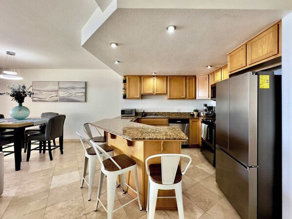 A modern kitchen with wooden cabinets, a stainless steel refrigerator, a granite countertop, and a dining area with bar stools.