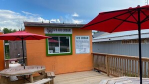 Un'area con decking in legno, tavoli da picnic e ombrelloni rossi, un cartello che indica 'FOOT'S ICE CREAM' e una bacheca con i gusti e i prezzi del gelato.