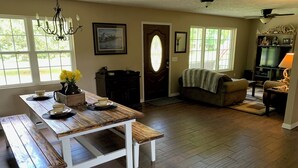 A dining area with a wooden table and benches, a chandelier, and a view of the outdoors through large windows.