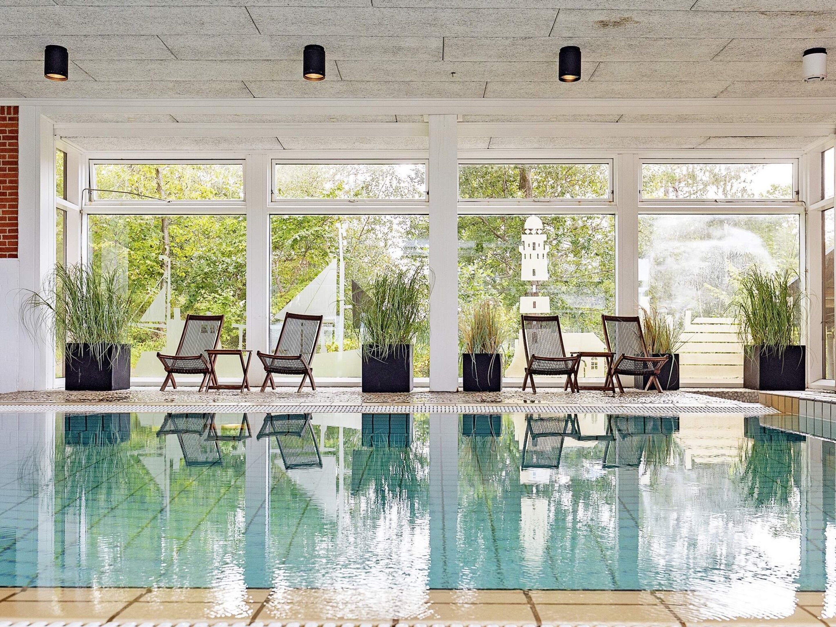 Una piscina interior con paredes de vidrio transparente, rodeada de plantas en macetas y sillas de mimbre.