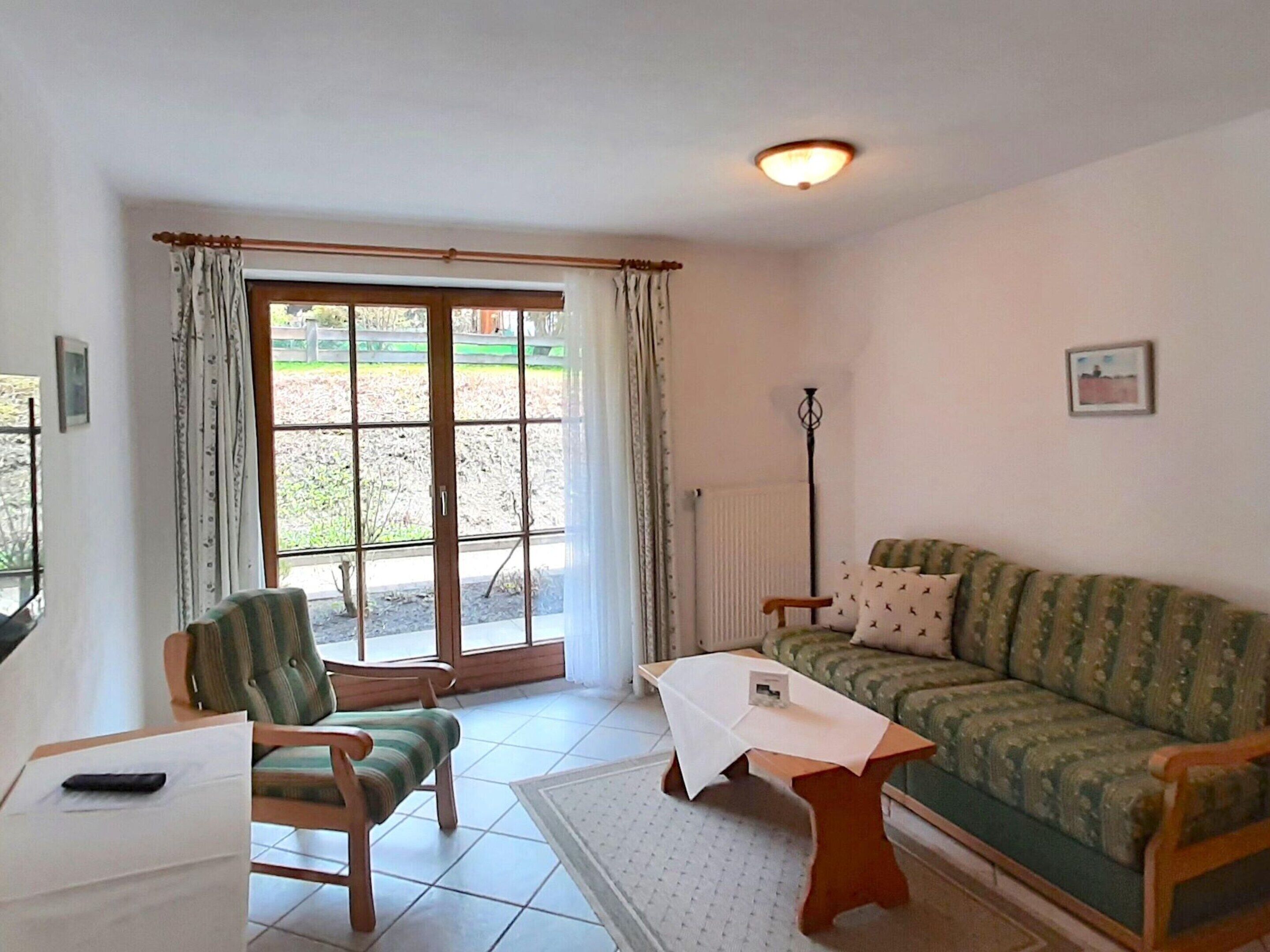 Sala de estar com sofá, duas poltronas, uma mesa pequena e vista para área externa através de uma porta de vidro.
