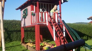 A red and white playhouse with a slide, surrounded by a wooden fence and a grassy area. - Holiday Apartment Tulpe Schuniglhof (Neukirchen Beim Heiligen Blut)