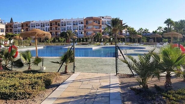 A resort with a swimming pool, palm trees, and a row of multi-story buildings with balconies.