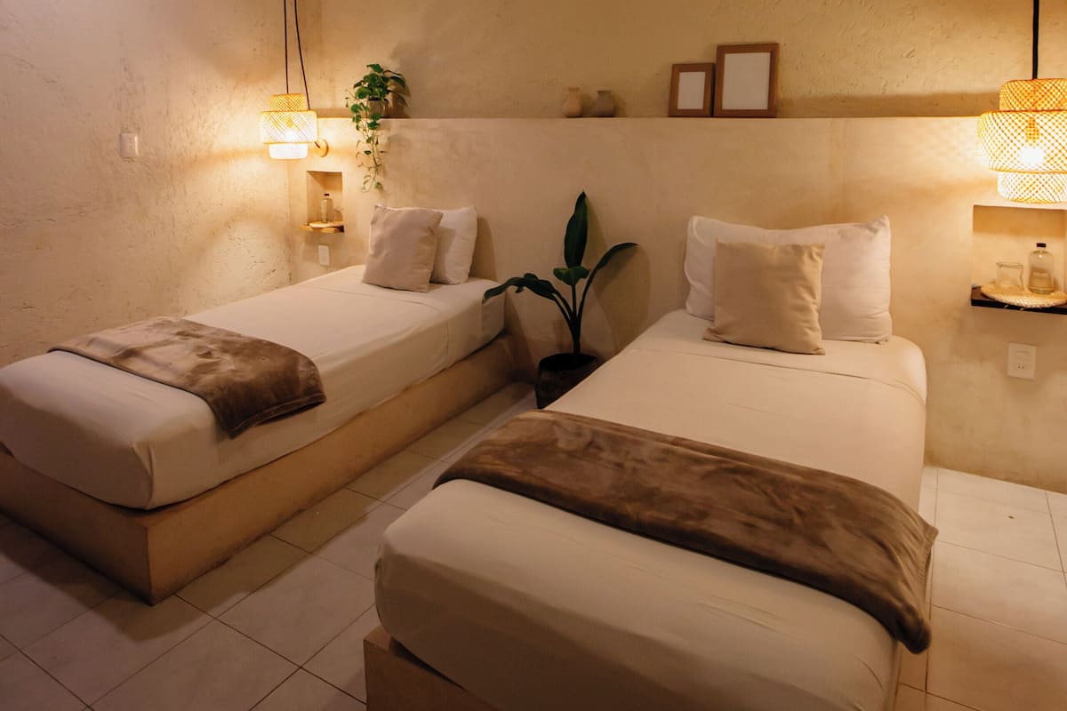 A hotel room with two single beds, a ceiling fan, wall-mounted lamps, and a small shelf with decorative items.