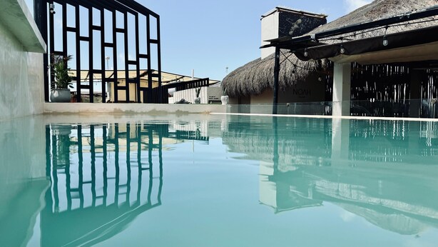 Piscina límpida refletindo a arquitetura ao redor, incluindo um edifício com telhado de palha e uma construção moderna com fachada de vidro.