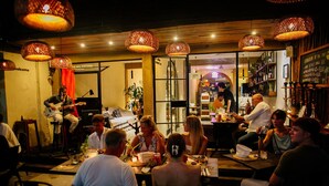Uma área de refeições ao ar livre com luminárias de vime, um palco com um guitarrista e um bar com cardápio em lousa.