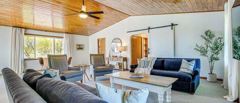 A spacious living room with a high wooden ceiling, a large window with curtains, a blue sofa, a coffee table, and a potted plant.