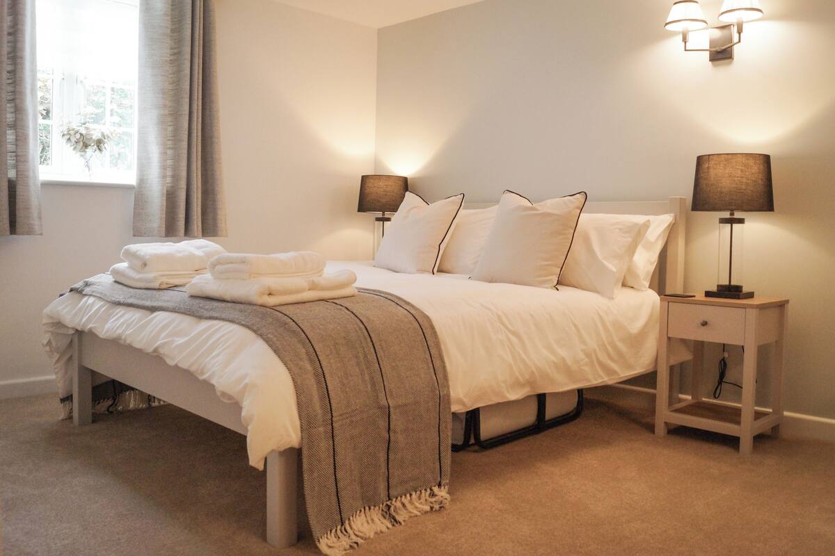 A neatly made bed with a white comforter and pillows, a bedside table with a lamp, and a window with curtains.