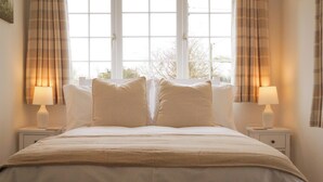 A neatly made bed with a beige bedspread and pillows, flanked by two bedside tables with lamps, in a room with large windows and curtains. - Lemon Tree (Dorchester)