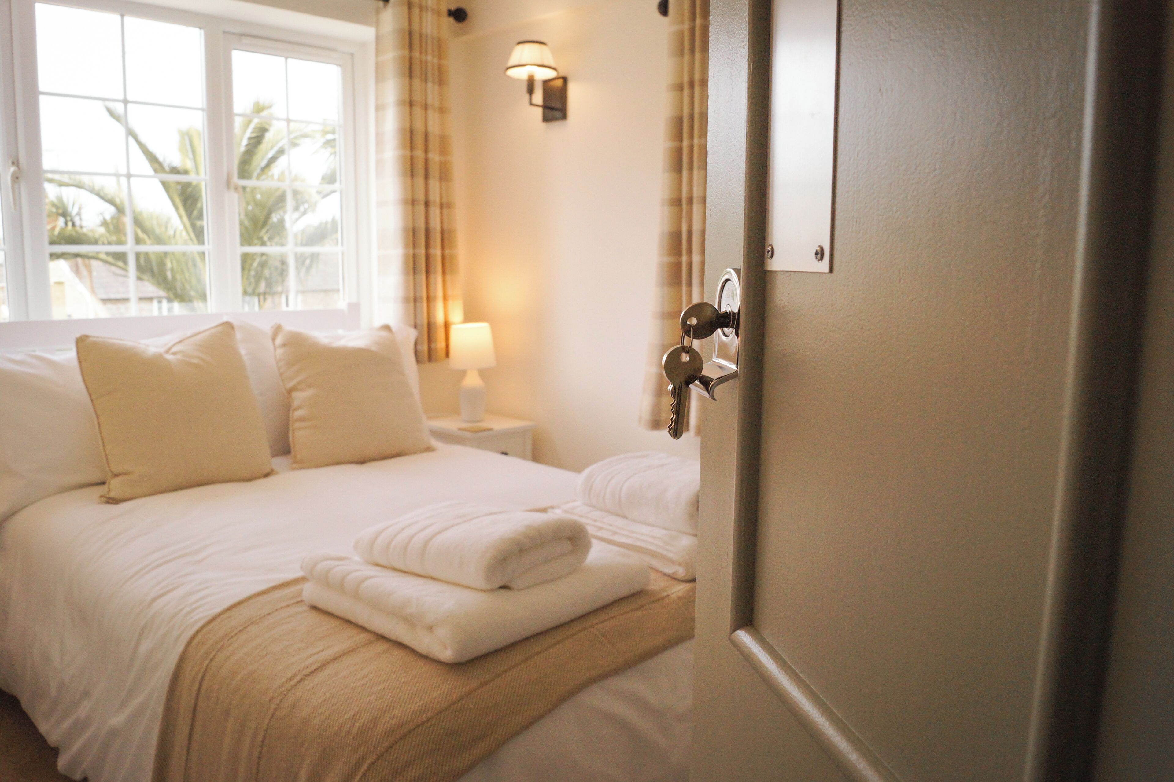 Habitación de hotel con cama, mesita de noche con lámpara y vista a palmeras a través de la ventana.
