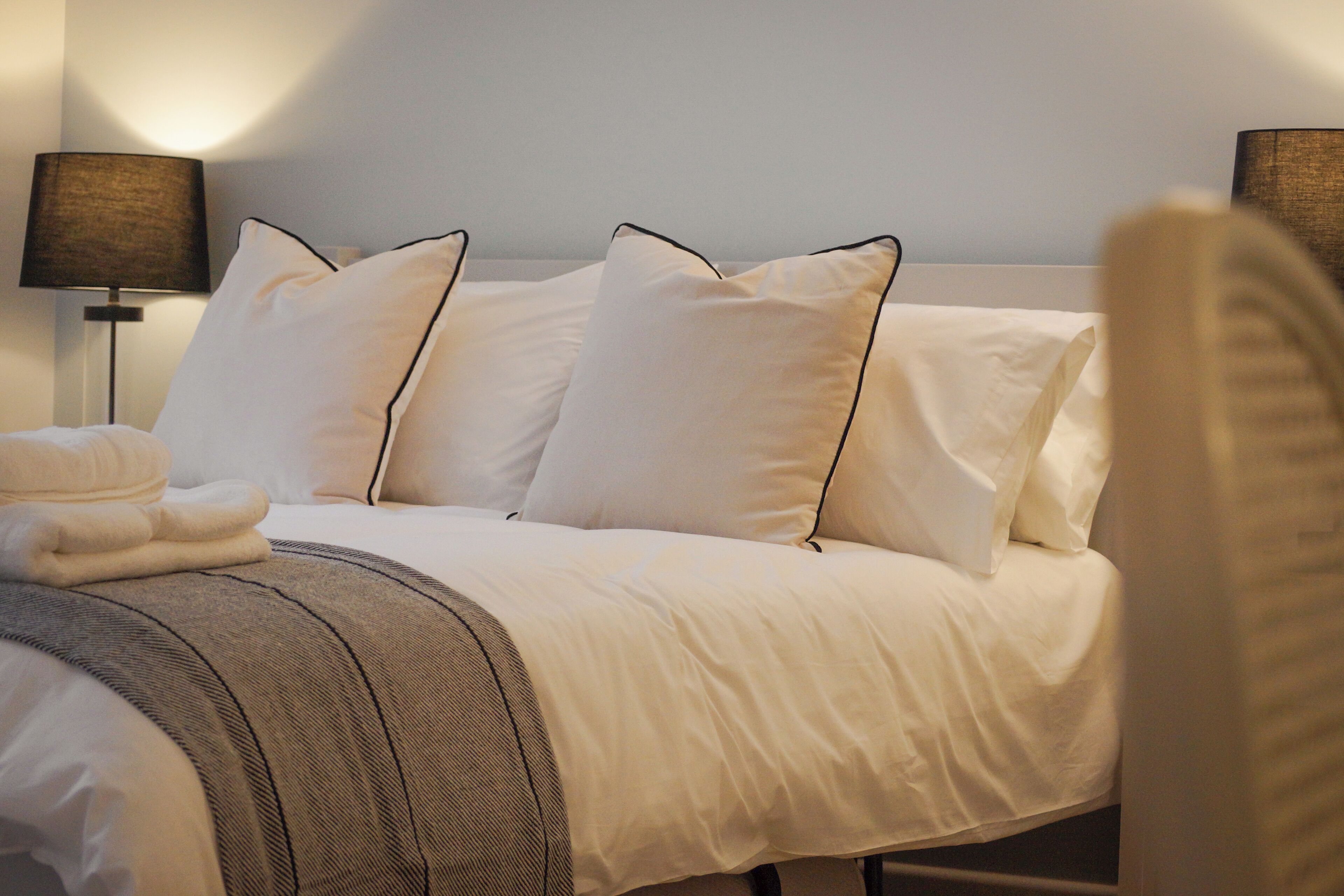 A neatly made bed with white linens and a grey throw blanket, two bedside lamps, and a folded towel on the side.