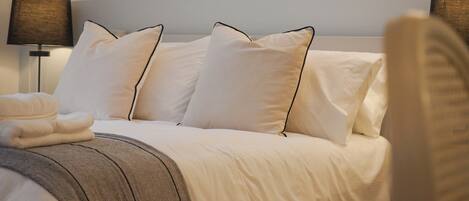 A neatly made bed with white linens and a grey throw blanket, two bedside lamps, and a folded towel on the side.