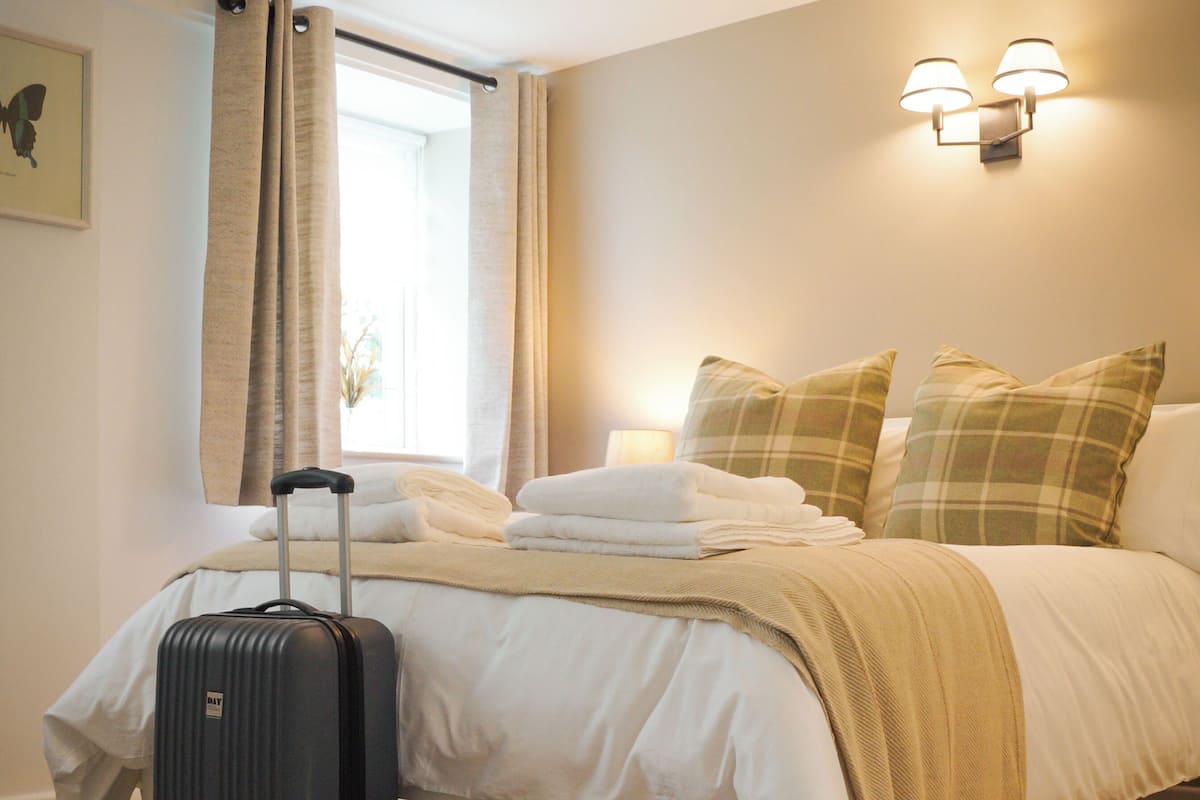 A neatly made bed with a suitcase, a window with curtains, and a wall-mounted light fixture.