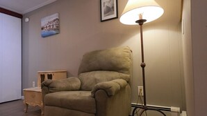 Uma sala de estar com uma poltrona bege, um abajur, uma pequena mesa lateral de madeira e um quadro pendurado na parede.