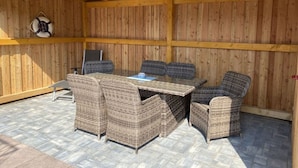 Une terrasse extérieure avec une table en bois et des chaises en osier, un gilet de sauvetage accroché au mur et une chaise en métal.