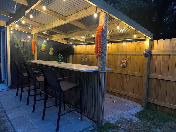 Un área de bar al aire libre con terraza de madera, taburetes de bar y una pantalla de televisión.
