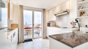 A modern kitchen with white cabinetry, a granite countertop, and a view of the beach through the sliding glass door. - Apartment Near Dunes in Koksijde (Koksijde)
