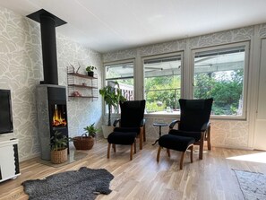 A cozy living room with a fireplace, two black armchairs, a small table, and a view of greenery through large windows. - 4 Star Holiday Home in Vastervik (Vastervik)