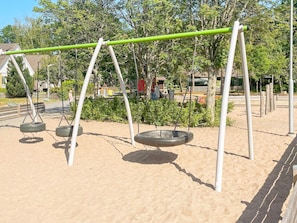 Ein Spielplatz mit Schaukeln und einer Gummireifenschaukel, umgeben von Sand und Bäumen.