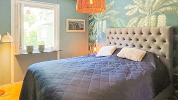A bedroom with a large bed, a window with blinds, a hanging orange light fixture, and a wall with a leafy pattern.