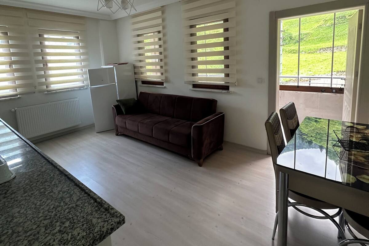 A spacious kitchen with a granite countertop, a dark brown sofa, a dining table with chairs, and large windows with blinds.