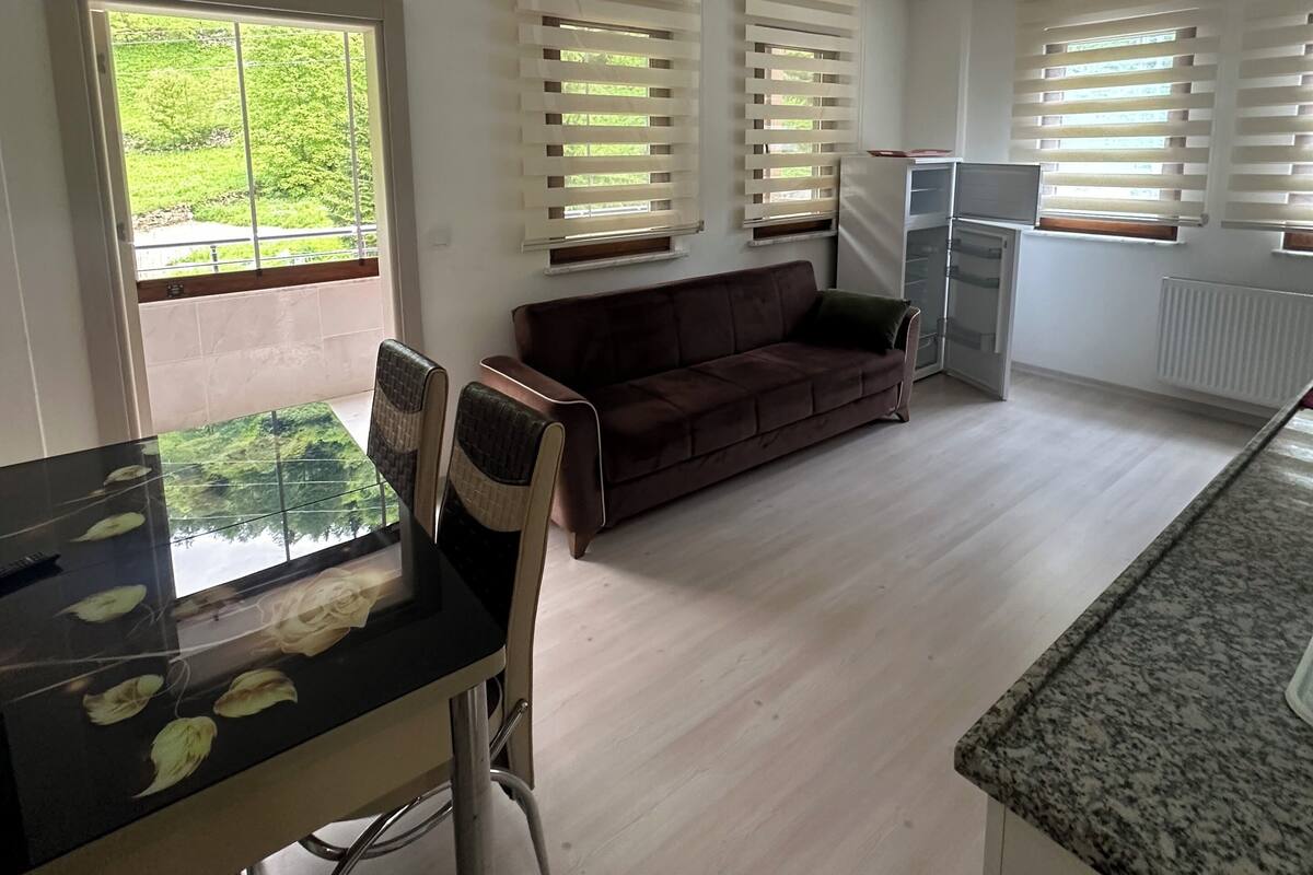 A modern kitchen with a dining area, a brown sofa, and large windows with blinds.