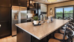 A modern kitchen with dark cabinets, a stainless steel refrigerator, a granite countertop, and a large island with chairs. - Luxury Chalet | Indoor pool | Hot Tub | Game Room (Sevierville)