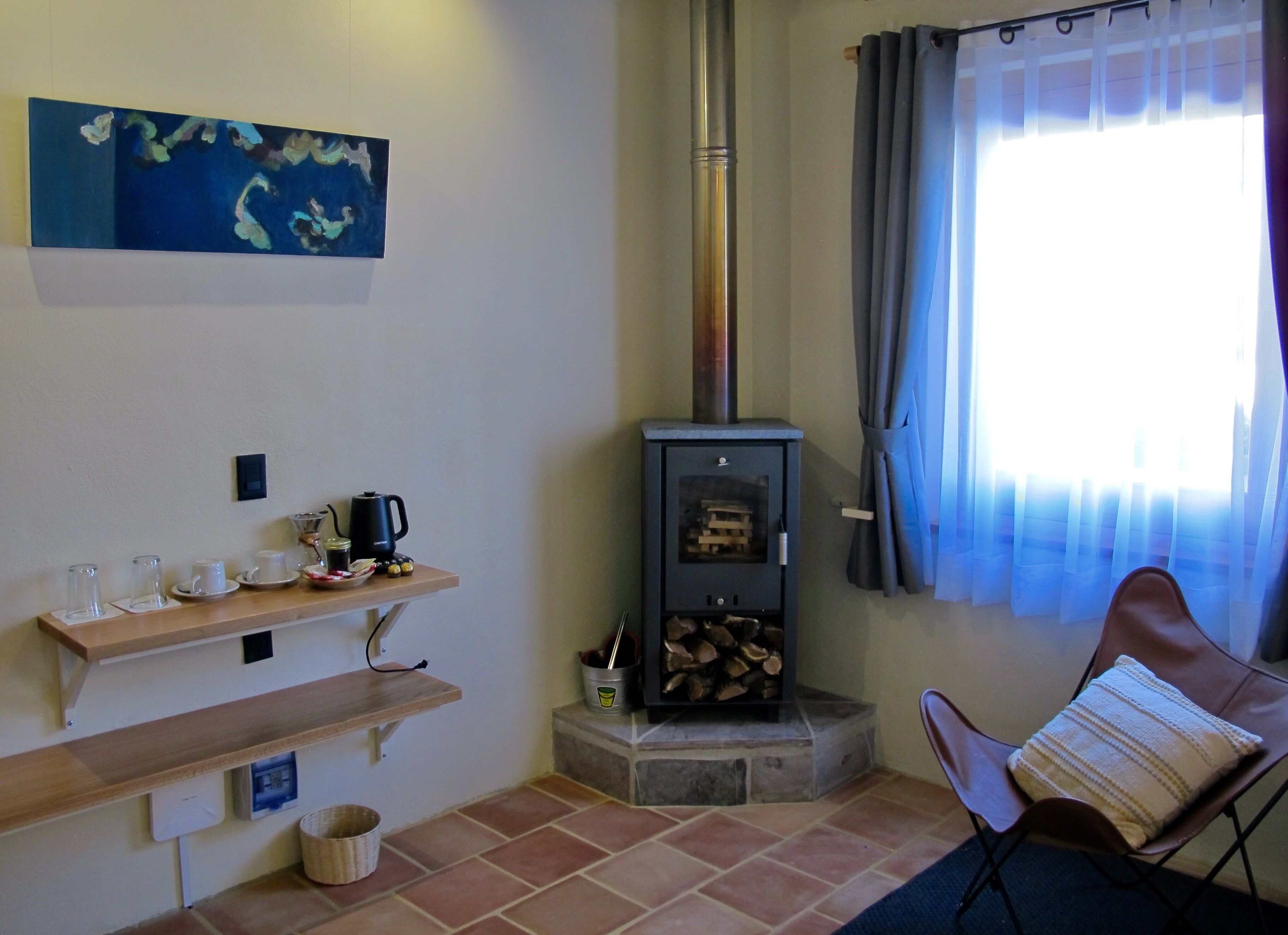 Una habitación acogedora con estufa de leña, una zona de café, un sillón con cojín y un cuadro en la pared.