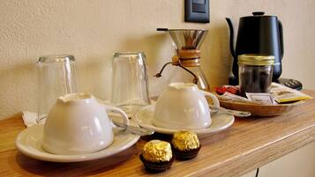 A breakfast setup with two white cups on saucers, a glass carafe, a coffee maker, a jar, and two wrapped chocolates on a wooden table.