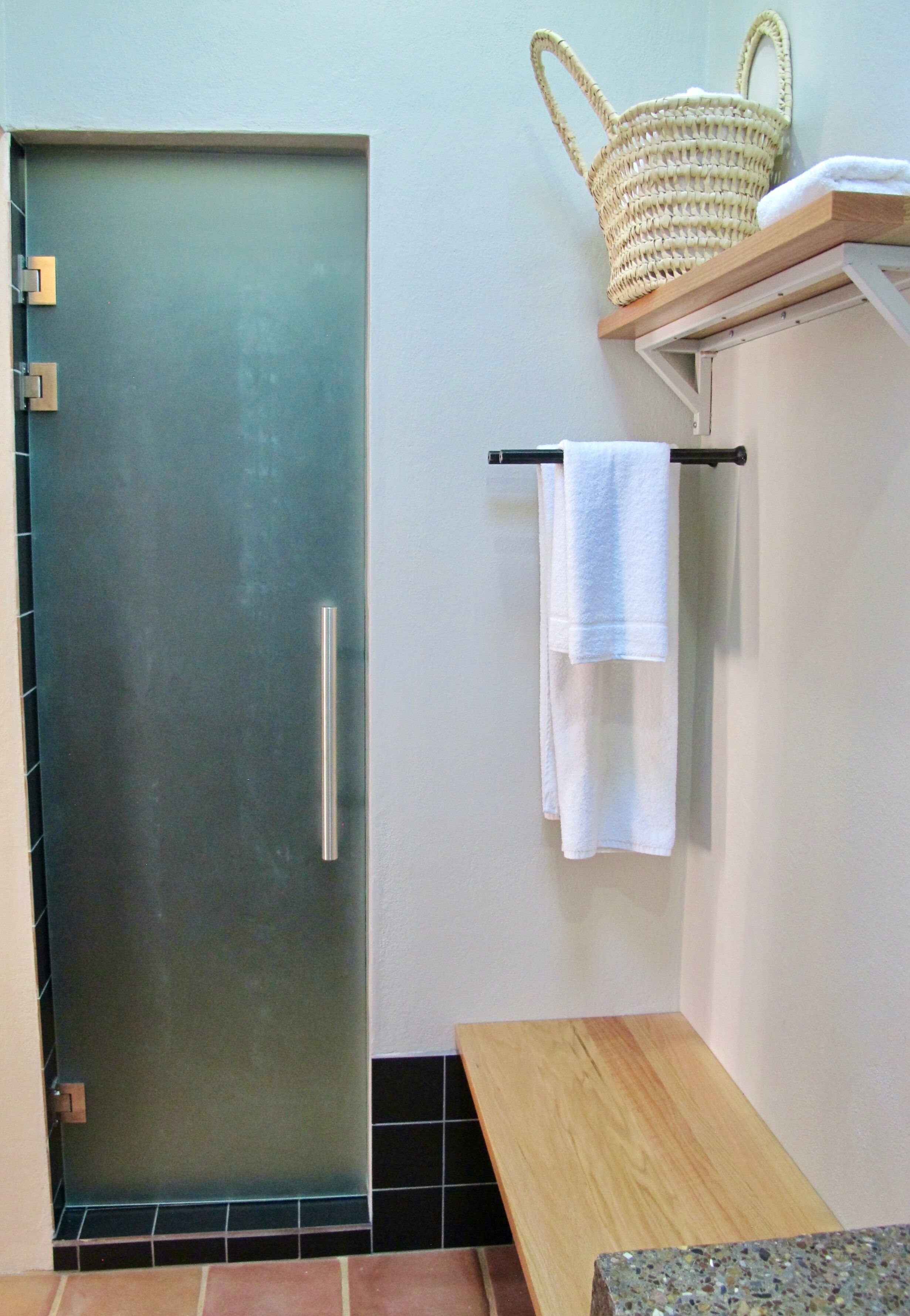 Un baño con puerta de vidrio satinado, un banco de madera, un perchero con una toalla blanca y una cesta tejida en un estante.