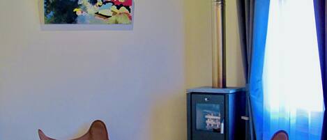 A living room with two chairs, a painting, a wood-burning stove, and a window with curtains.