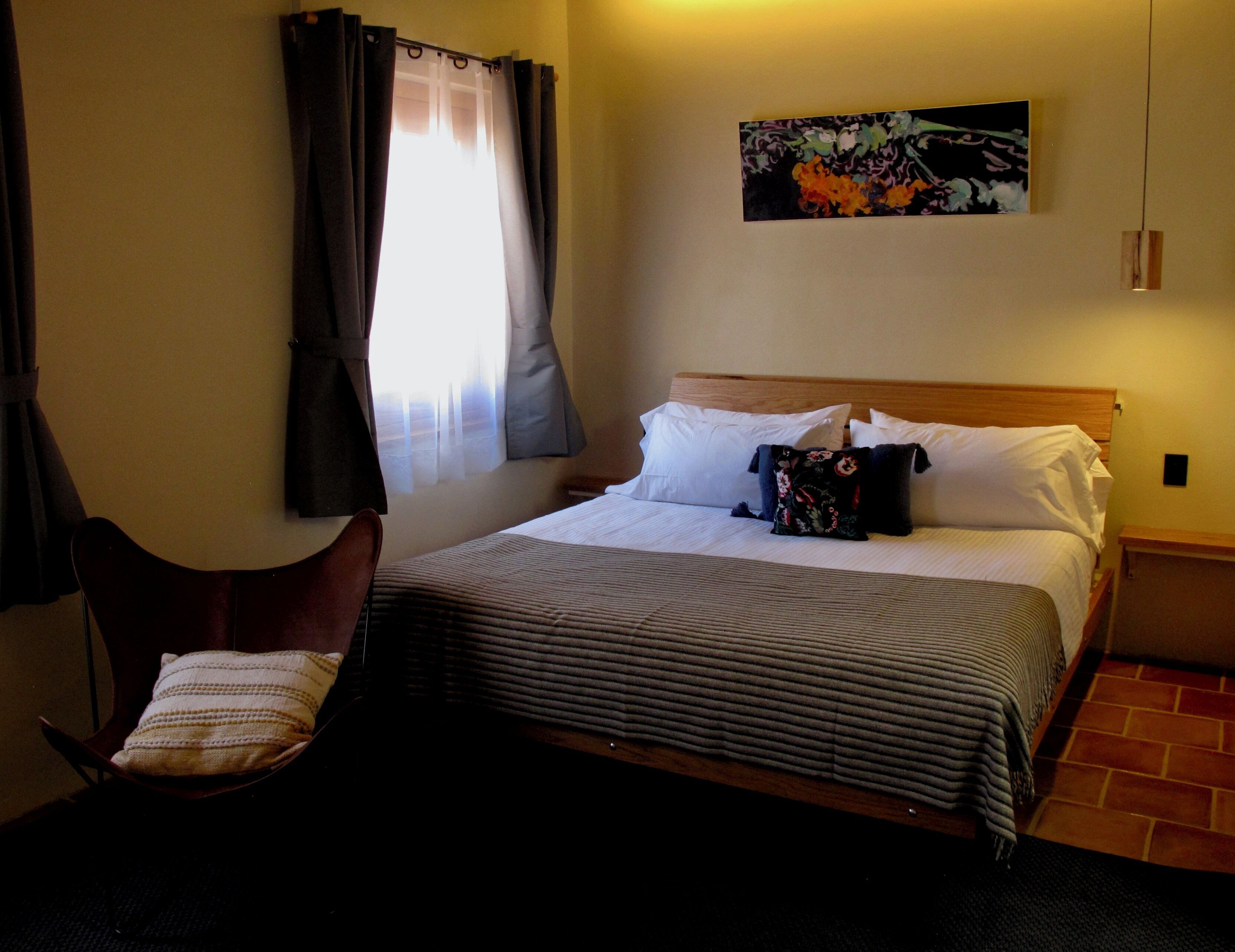 A hotel room with a bed, a chair, a painting on the wall, and a window with curtains.