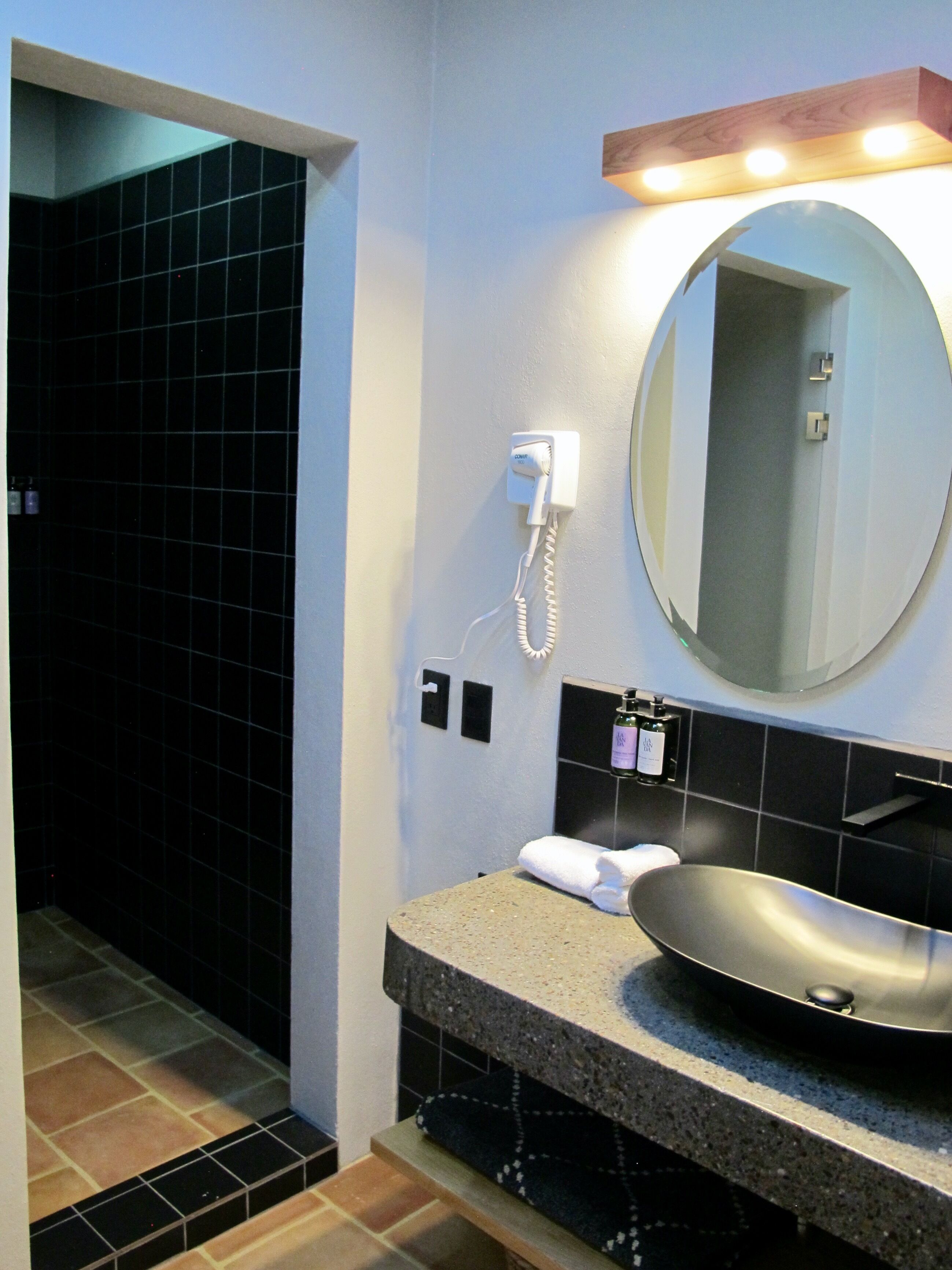 A bathroom with a black tiled shower, a sink with a round basin, a mirror with lights above, and a towel on the counter.
