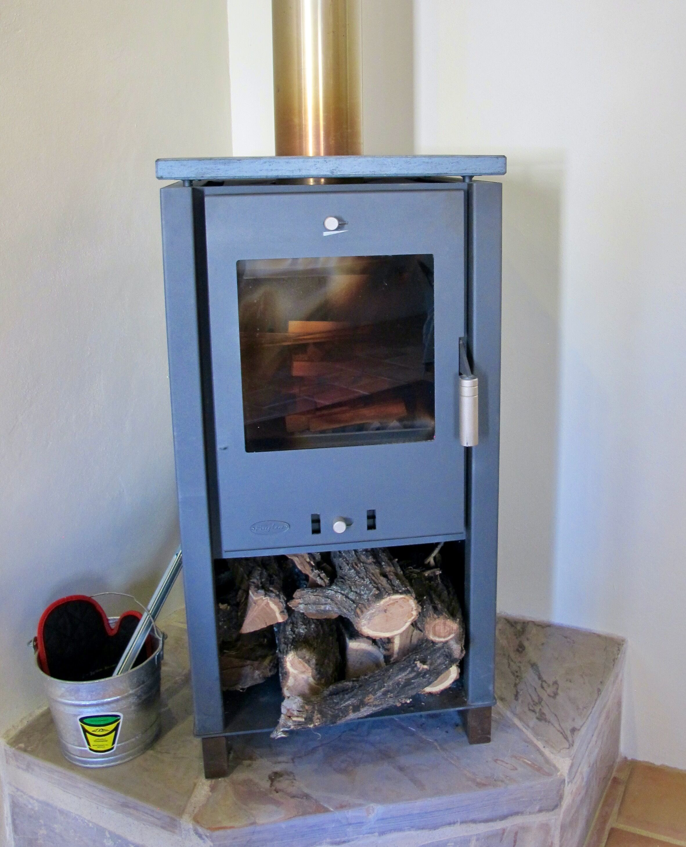 Un estufa de leña con puerta de vidrio, un balde de metal con tapa con forma de corazón y un compartimento para almacenar leña.