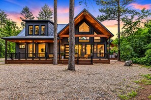 Une cabane en rondins moderne, dotée d’une grande terrasse et de nombreuses fenêtres, entourée d’arbres et de gravier.