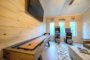 A cozy room with a foosball table, two arcade games, a bench, and a window with a view of trees.