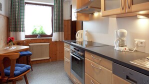 Ett kompakt kök med träskåp, en inbyggd ugn, en kaffebryggare och ett fönster med gardiner.