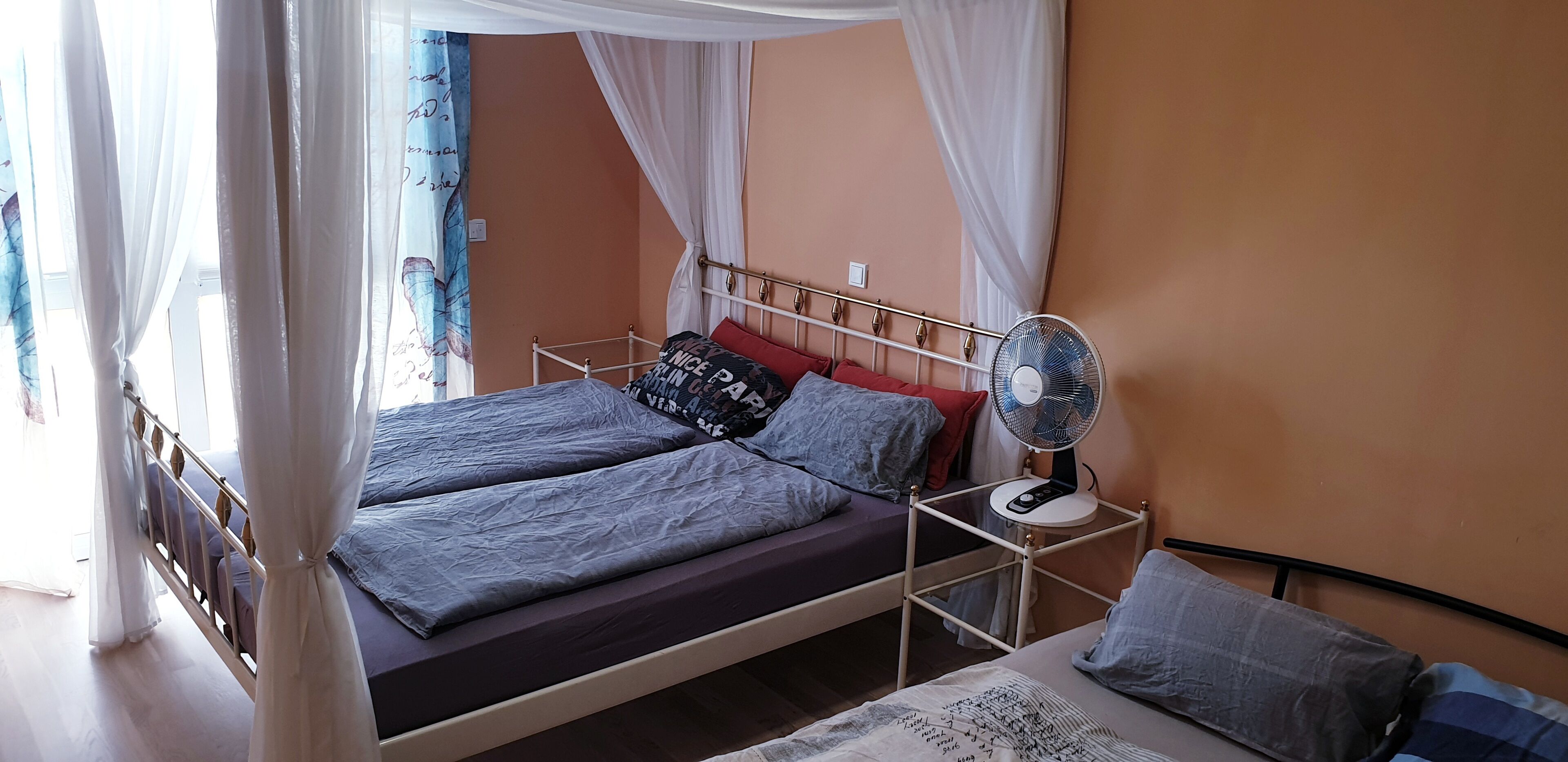 Ein Bett mit Baldachin, ein Nachttisch mit Ventilator und ein Fenster mit Vorhängen.